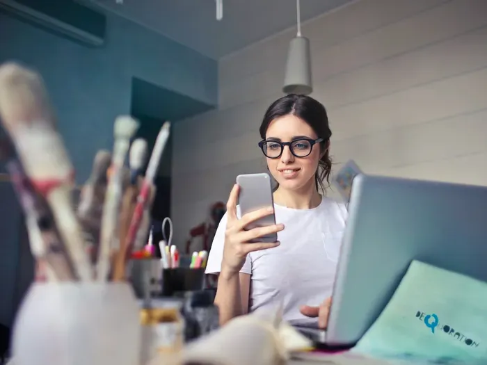 Small business owner checks phone at a desk with laptop and art supplies, updating a listing in a local directory